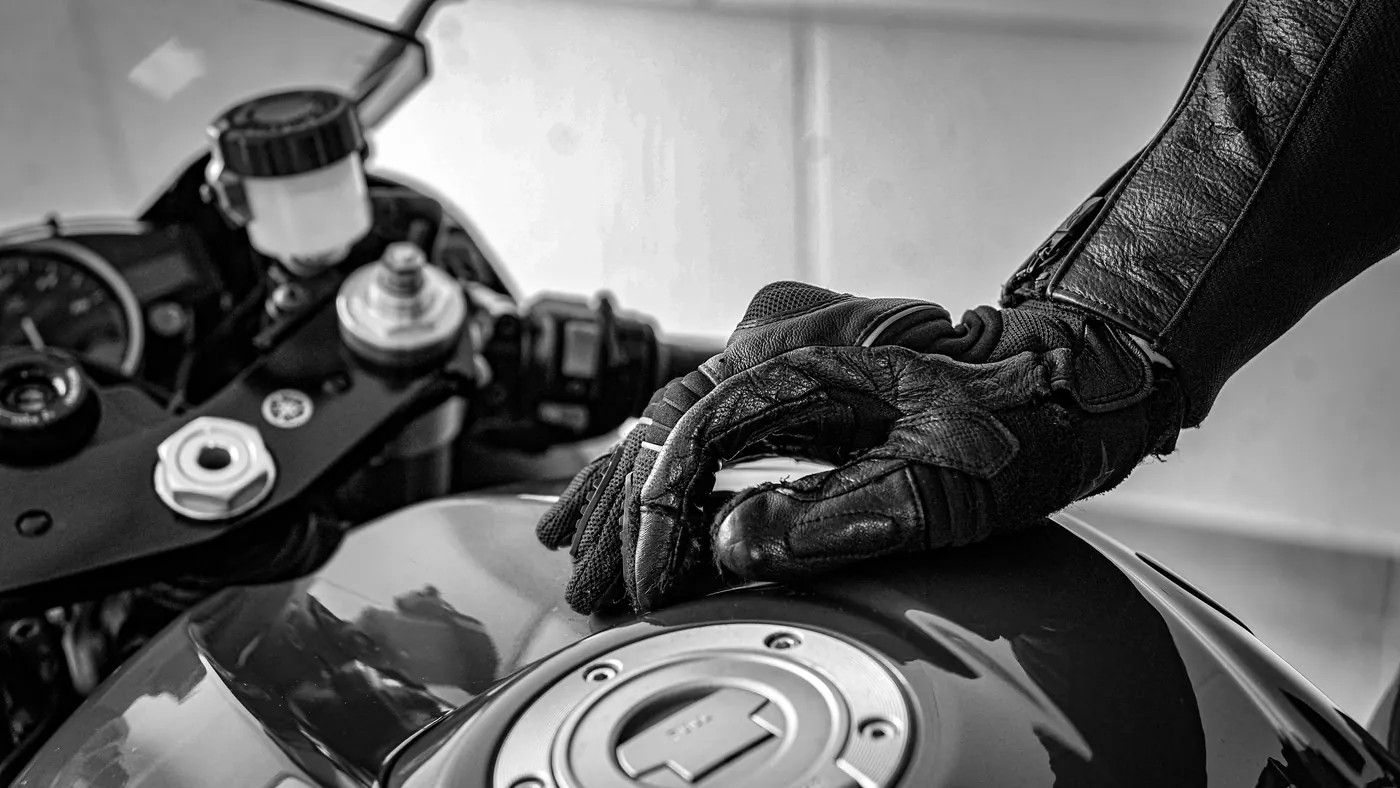 Gloved hand and leather jacket sleeve resting on a motorcycle fuel tank, black and white