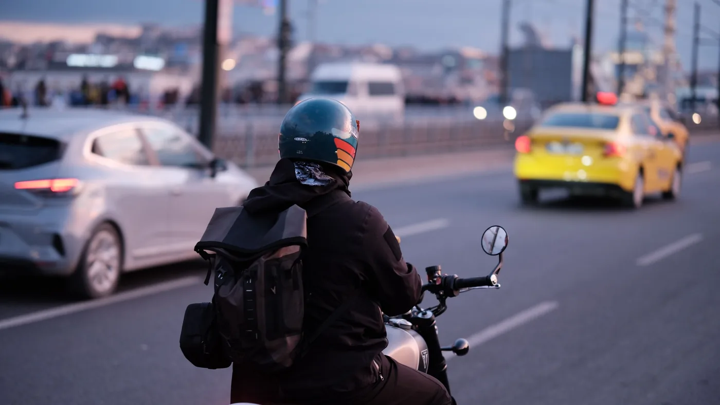 Motorcyclist with helmet and backpack riding in city traffic among cars at dusk, seen from behind