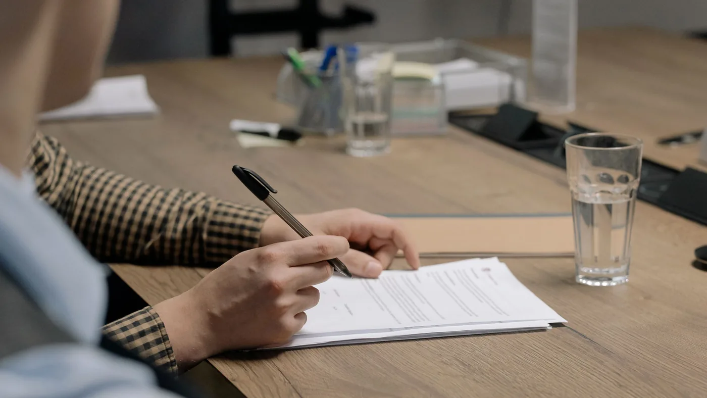 Person in a plaid jacket signing paperwork at a wooden office table