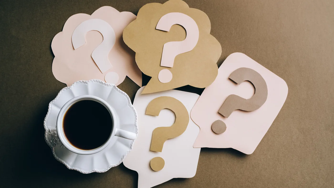 Paper cutouts of question marks next to a cup of black coffee on a brown table