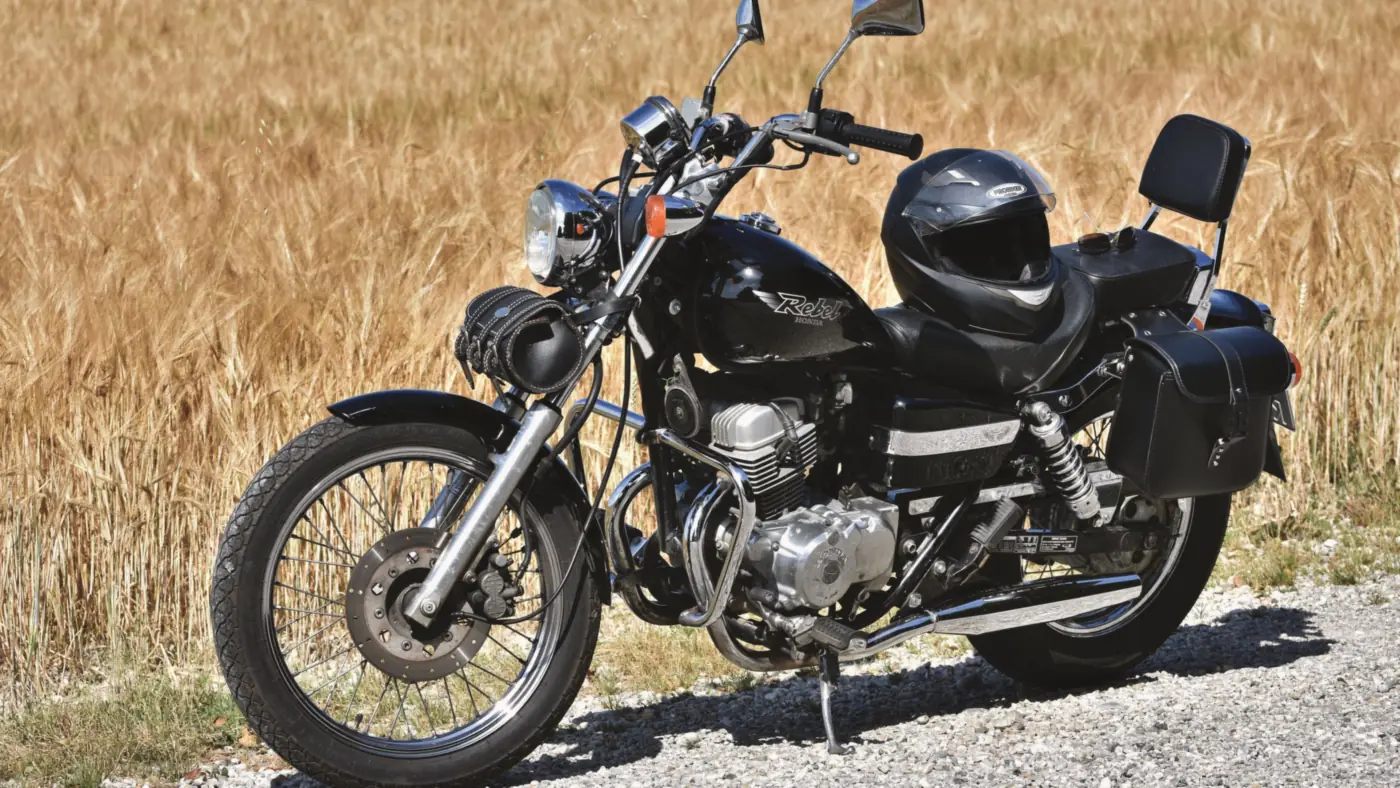 Black cruiser motorcycle with a helmet on the seat, parked on a gravel shoulder beside a wheat field