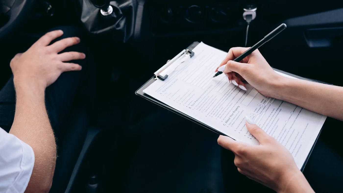 Hands filling out an insurance form on a clipboard inside a parked car