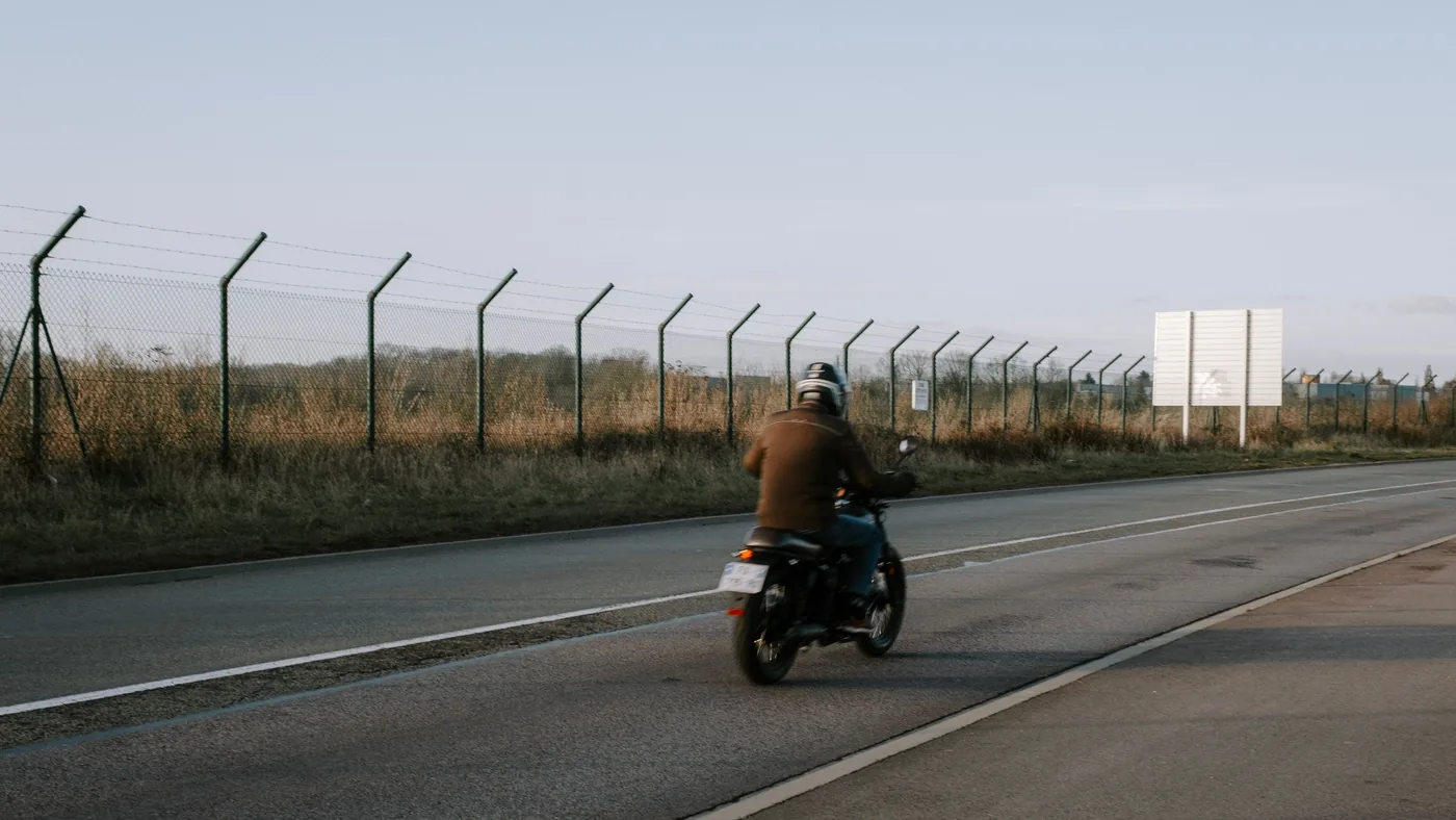 Motorcyclist in a jacket and helmet riding along a two-lane road beside a fenced grassy field, seen from behind